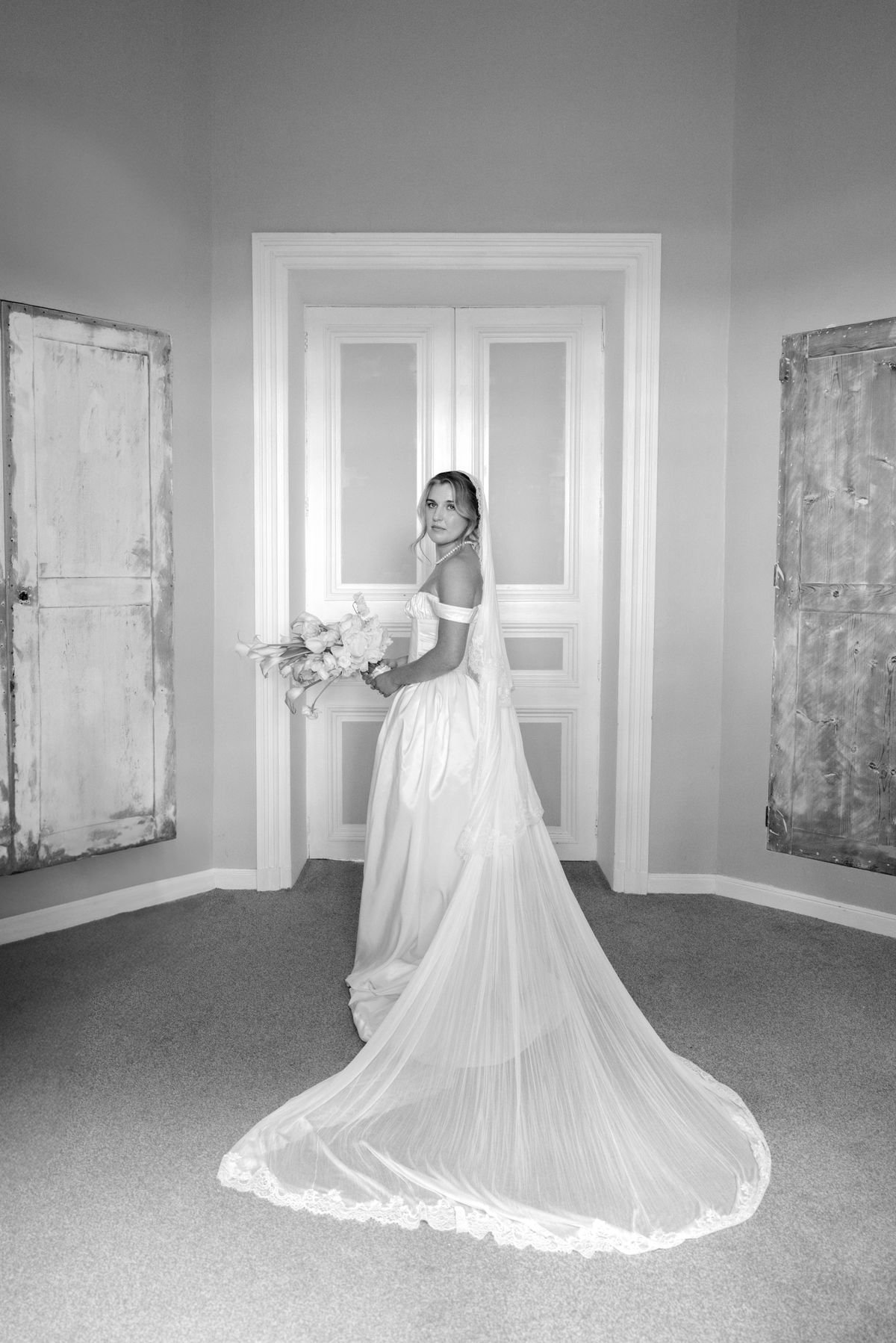 The bride in an off-the-shoulder gown and long veil looks back over her shoulder, holding her bouquet, in black and white.
