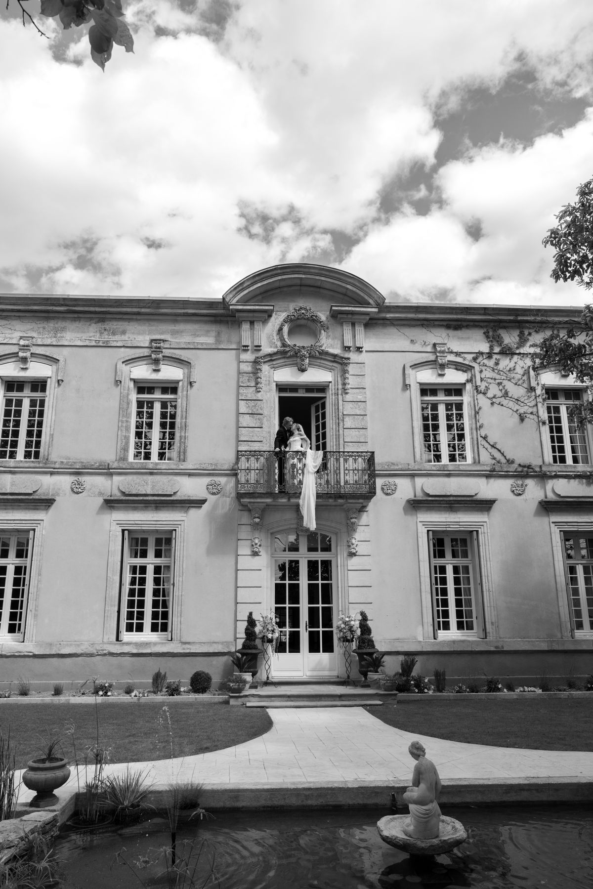 A couple stand close together on the balcony of a grand stone manor, the bride's long veil hanging down, in black and white.