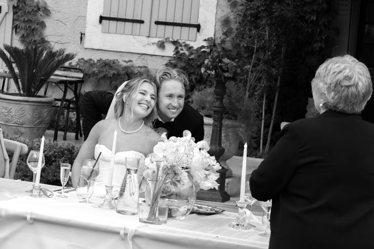 The bride laughs at a candlelit table with flowers as a guest leans in beside her and an older woman stands in the foreground.