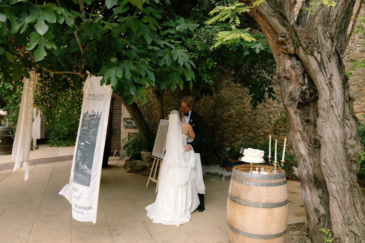 The bride in a long veil and the groom embrace beneath a large tree, near a signboard and a cake on a wine barrel.