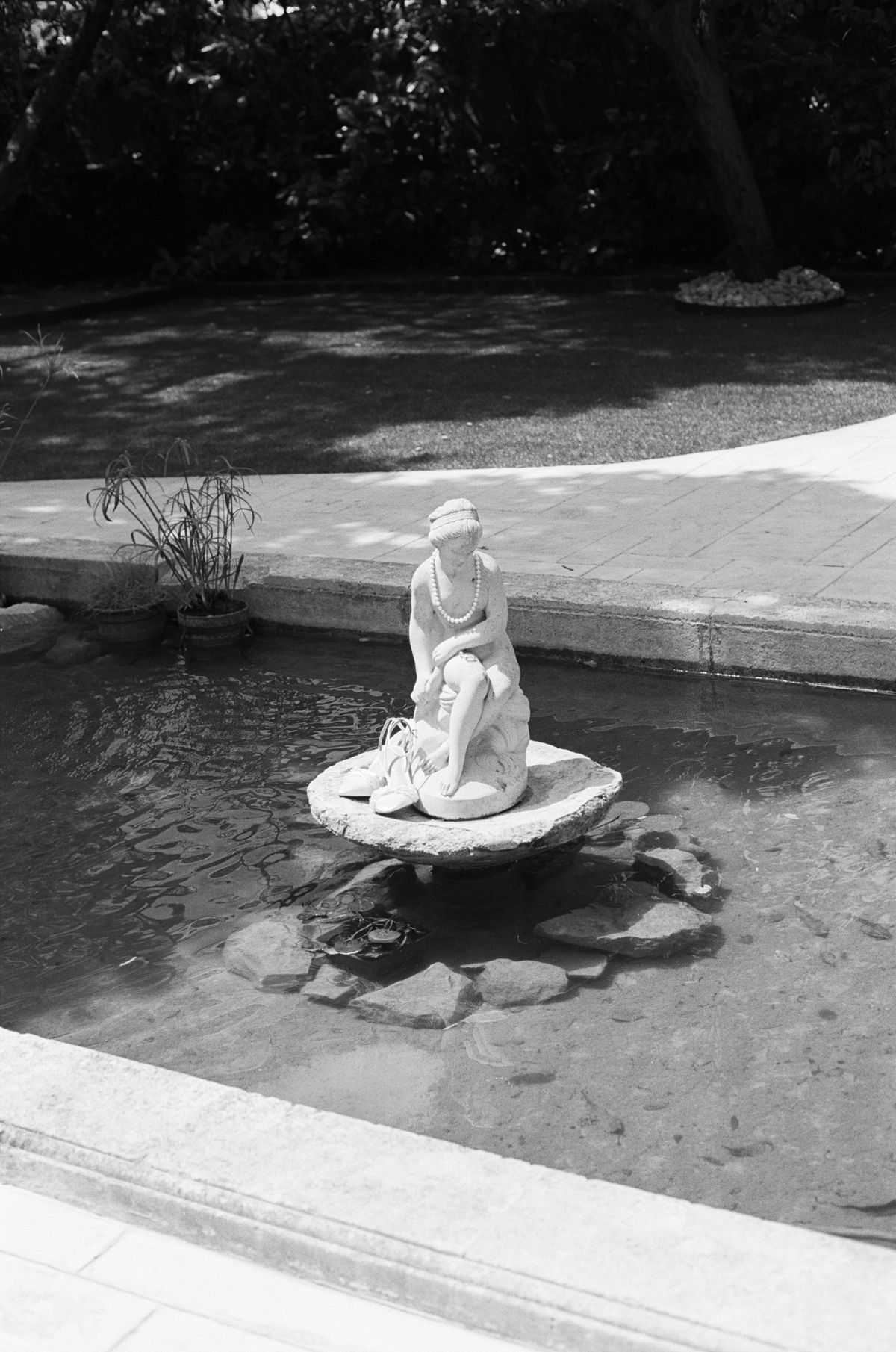 In black and white, a white statue of a seated figure stands on a small pedestal in the middle of a still garden pool.