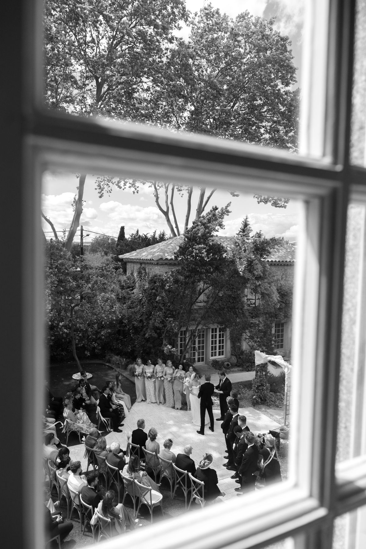 In black and white, a window frames seated guests and a wedding party gathered for an outdoor ceremony under trees.