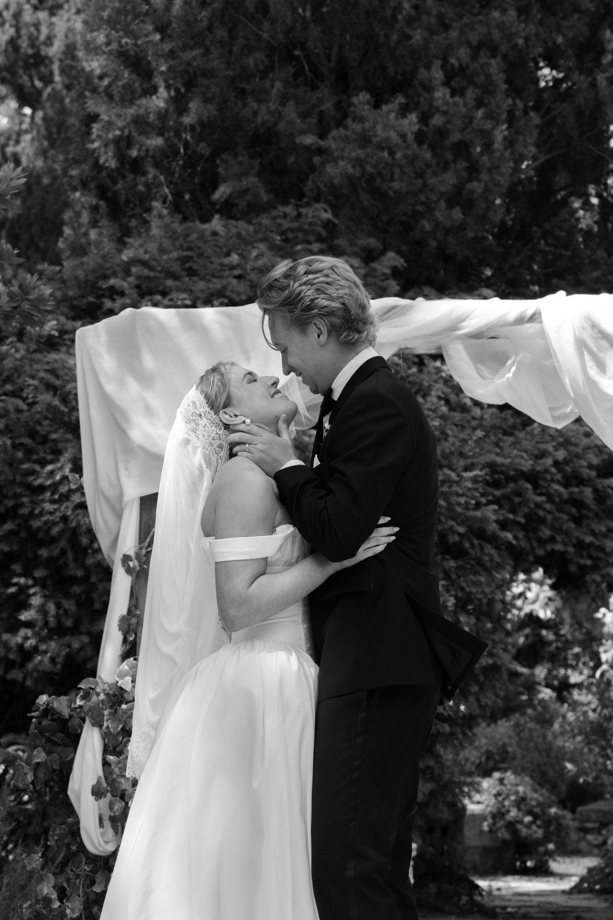The couple embrace and kiss in a garden beneath a draped white canopy, the bride wearing a veil.