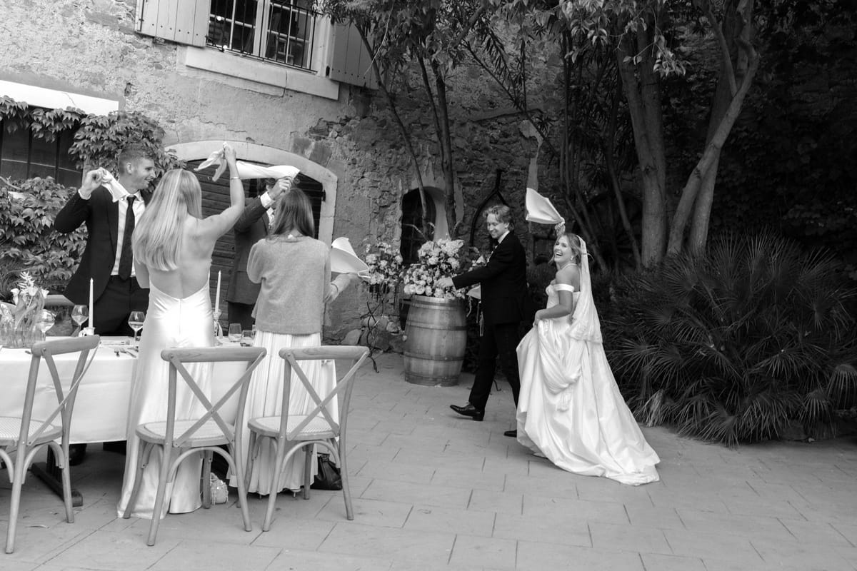 In black and white, the bride walks across a courtyard past set tables while guests wave napkins in the air.