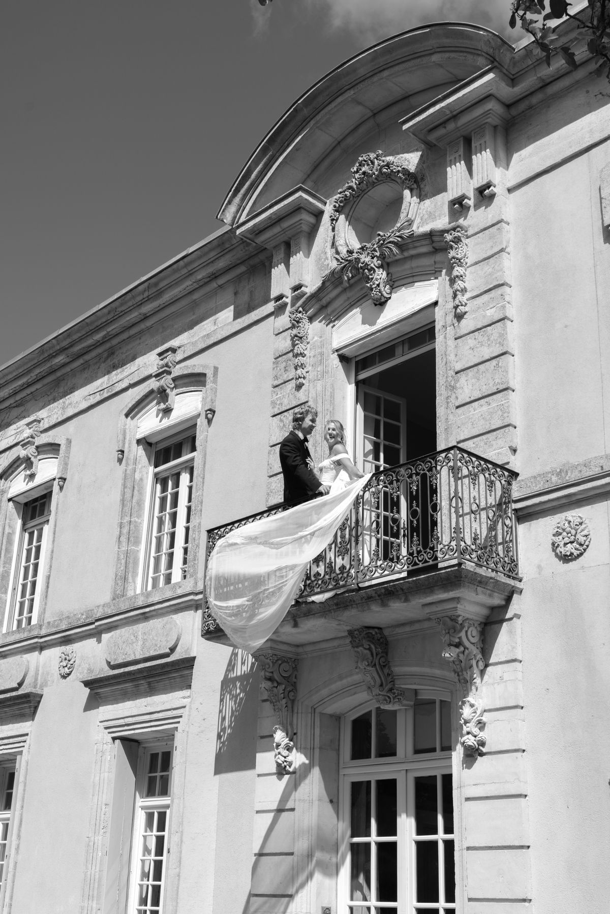 The couple stand together on an ornate wrought-iron balcony of a stone château, the bride's long veil streaming below.