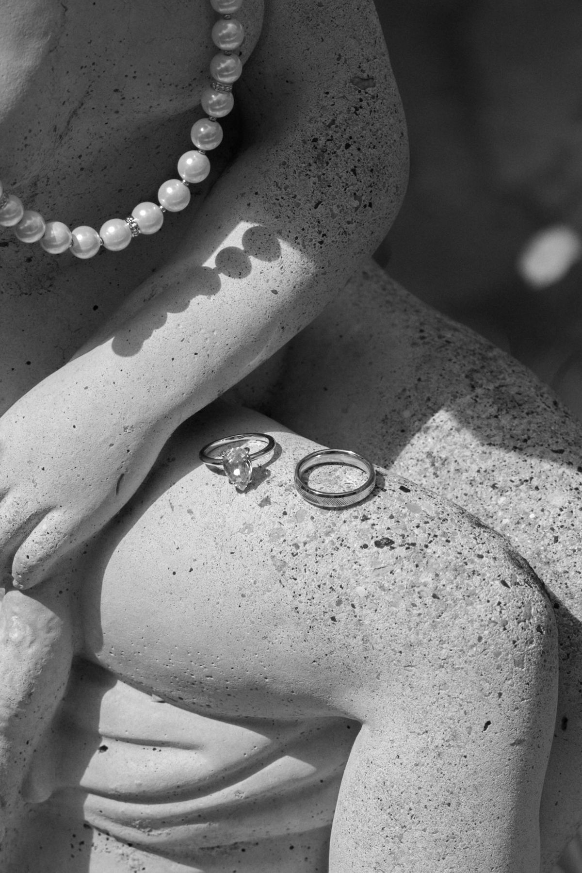 Two wedding rings sit on the stone hand of a statue, beside a string of pearls draped over it.