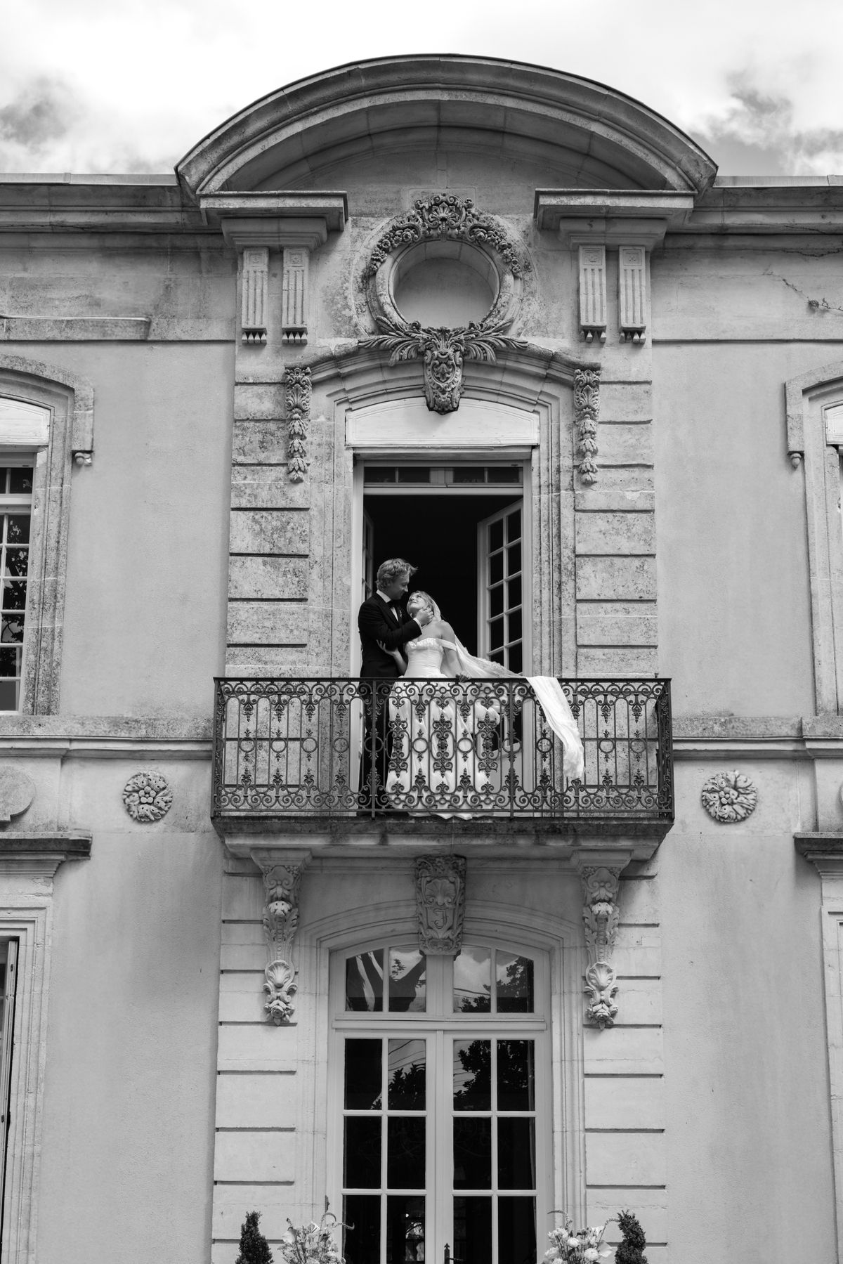 The couple stand together on a wrought-iron balcony beneath an arched pediment of a stone chateau under a cloudy sky.