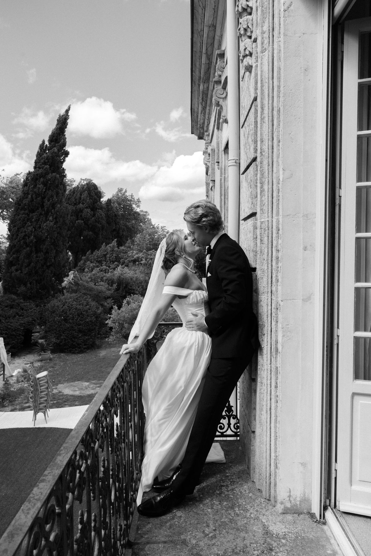 In black and white, the couple kiss on a balcony beside tall French windows, with trees and a stone balustrade around them.
