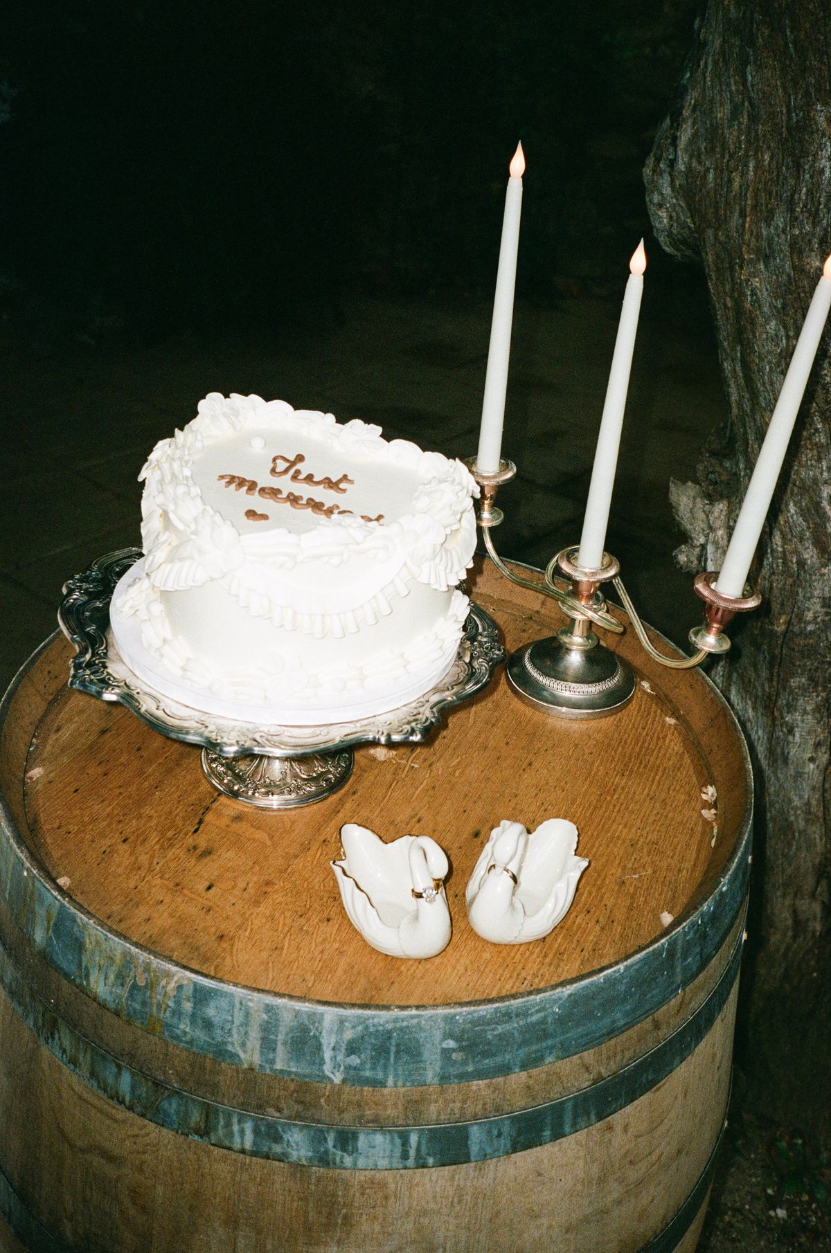 A small white heart-shaped cake with script lettering sits on a wooden barrel beside three tall candles and two white ceramic swans at night.