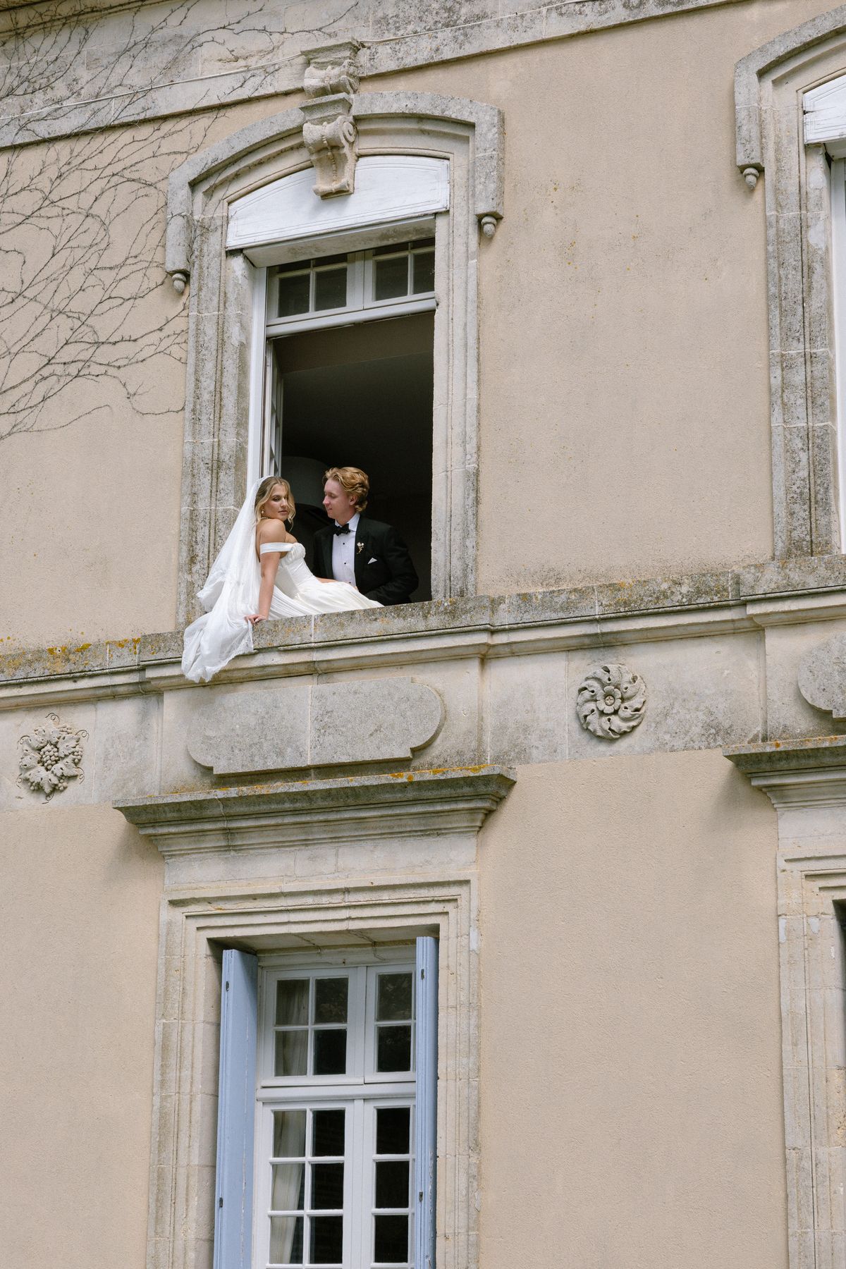 The bride, seated on a window ledge, and the groom lean together in an upper window of a stone chateau.