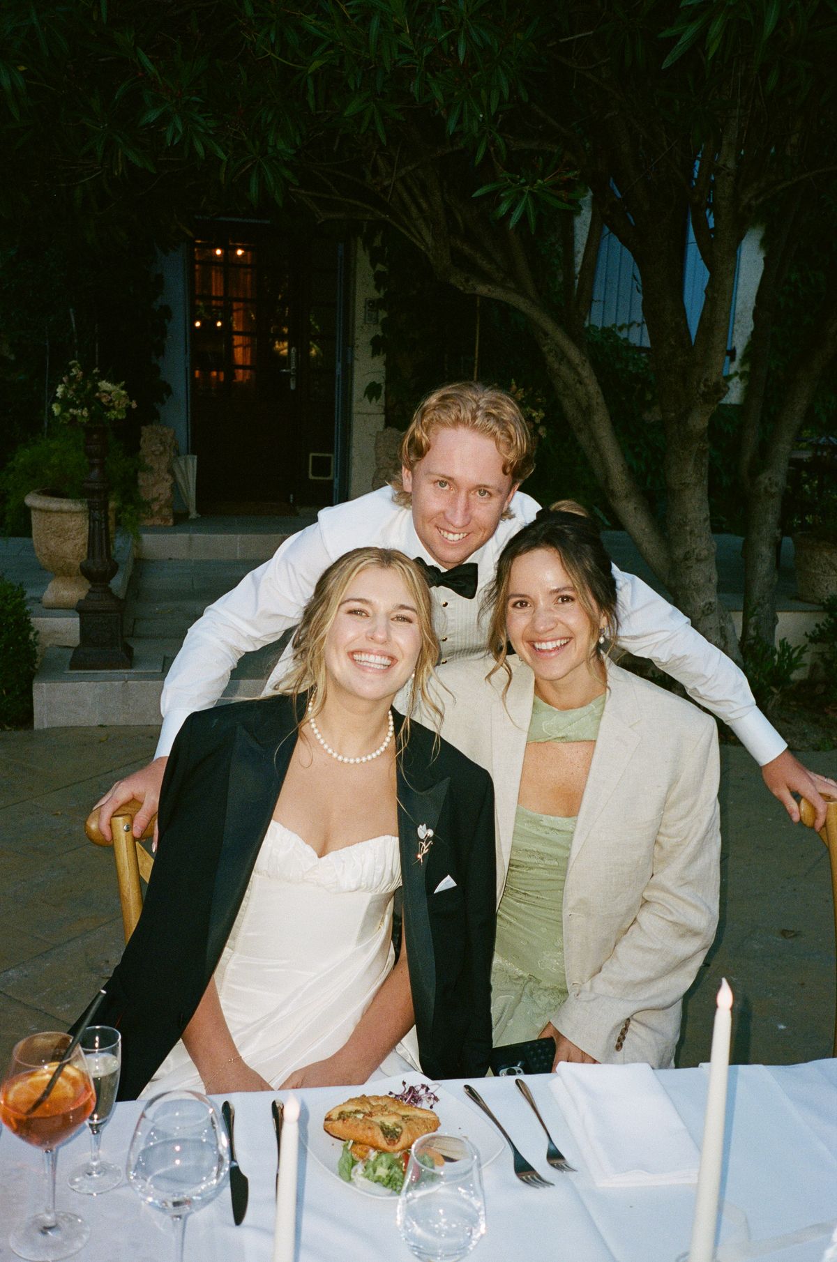 The bride in a black blazer sits at a dinner table smiling, with two people beside and behind her, a cocktail in front.