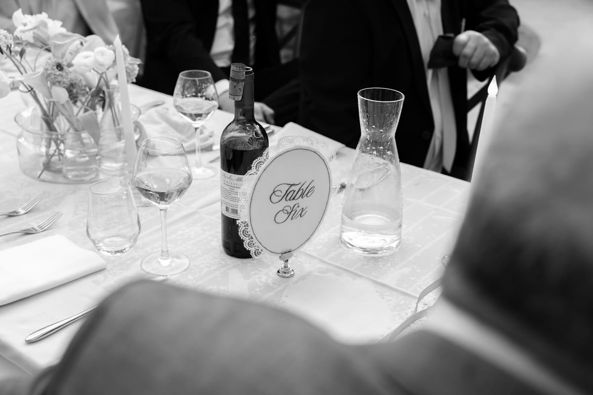 A table number card beside a bottle of red wine, glasses and a water carafe on a white dinner table.