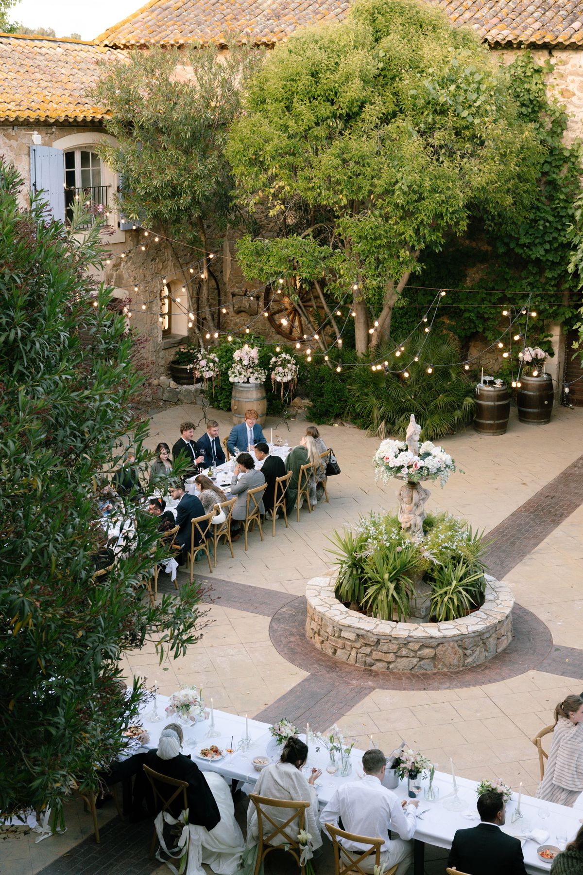 Seen from above, guests sit at long tables in a courtyard with a stone fountain, trees and string lights.