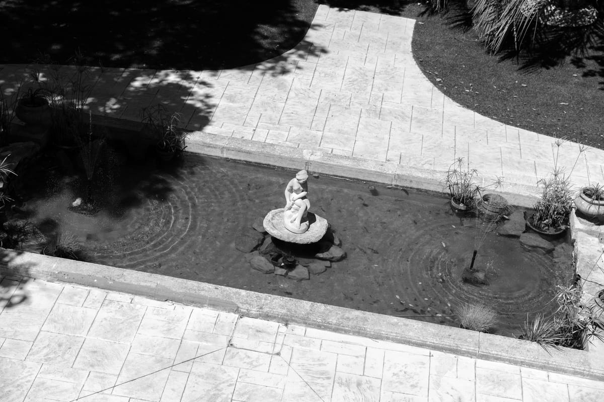 In black and white, a stone statue stands in a small pond beside a paved courtyard, seen from above.