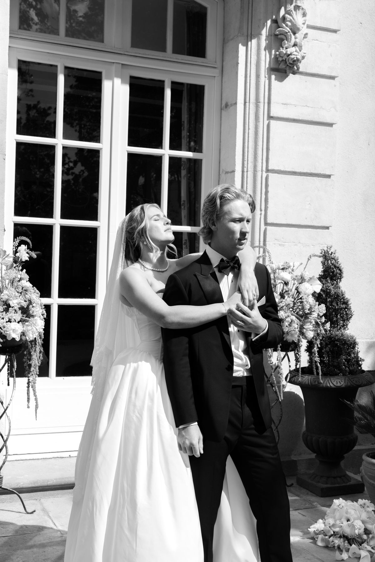 The bride in a veil and the groom in a dark suit embrace beside tall glazed doors, both looking away from the camera.