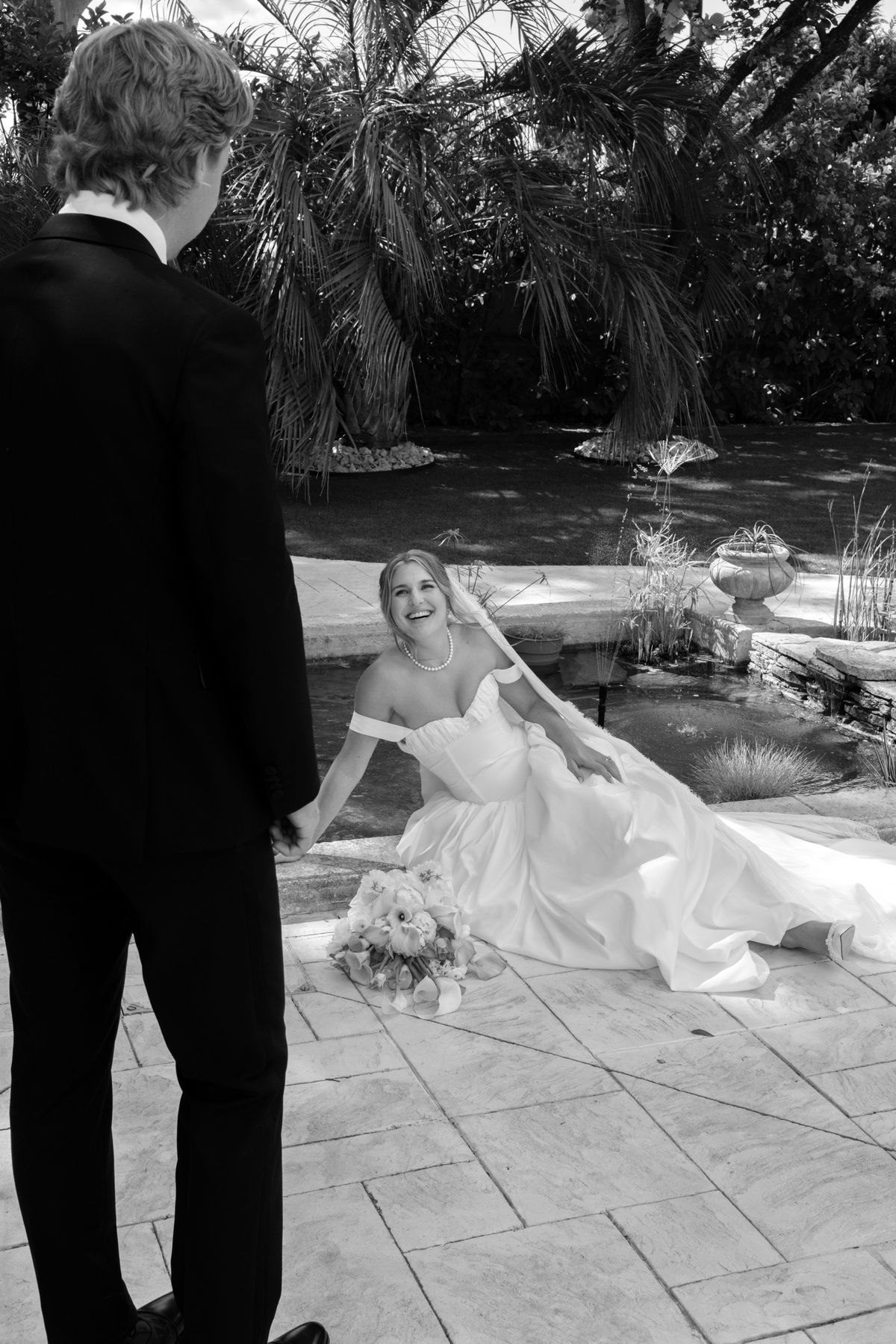 The groom stands in the foreground looking down at the bride, who sits in her gown beside a garden pond with palm trees behind.