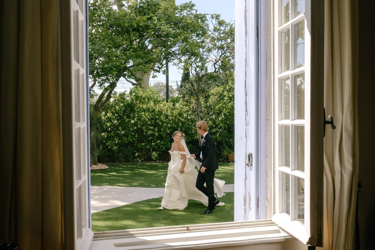 Seen through an open window, the couple walk hand in hand across a sunlit green lawn lined with trees.
