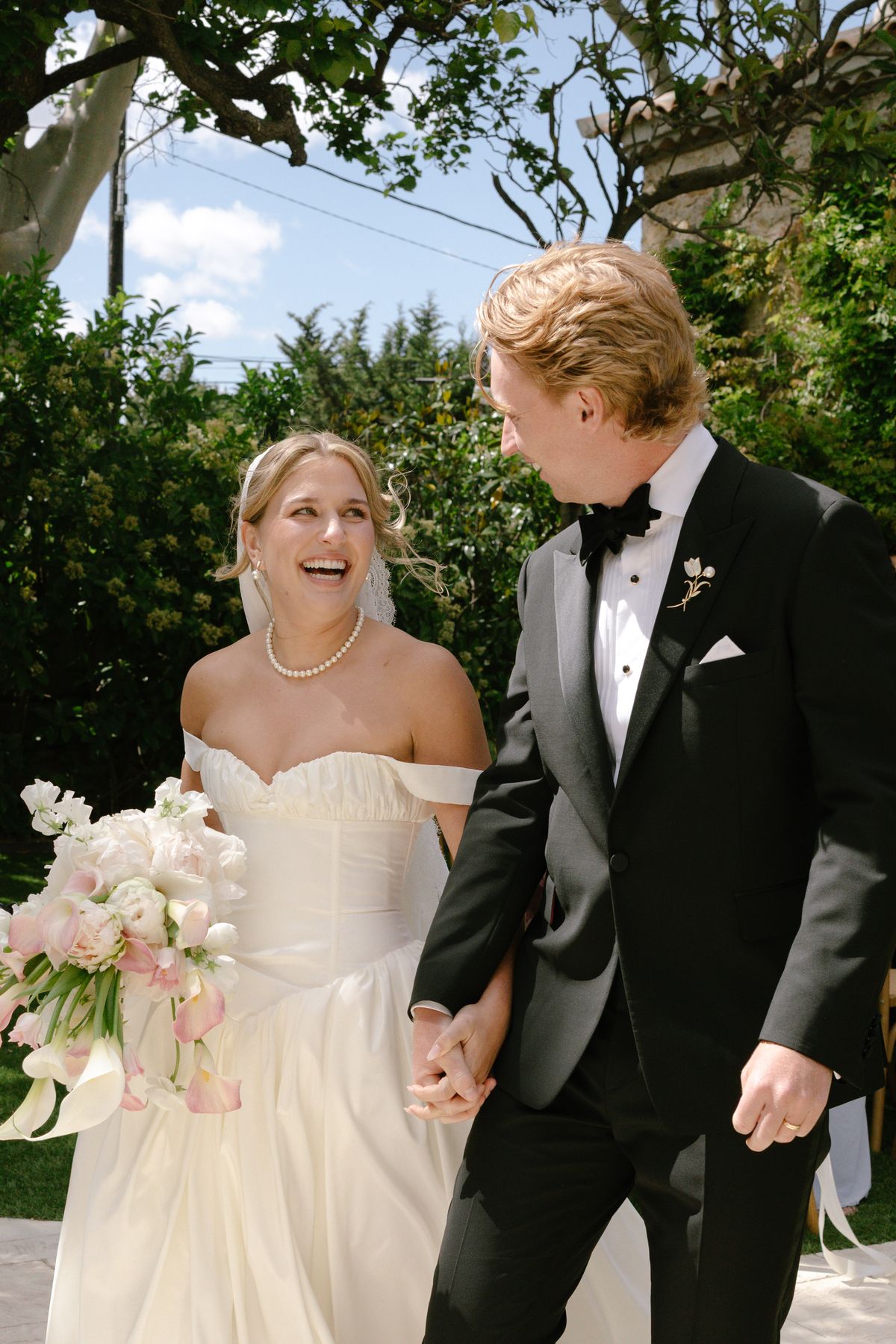The laughing bride in an off-the-shoulder ivory gown holds hands with the groom in a black tuxedo, in a leafy garden.