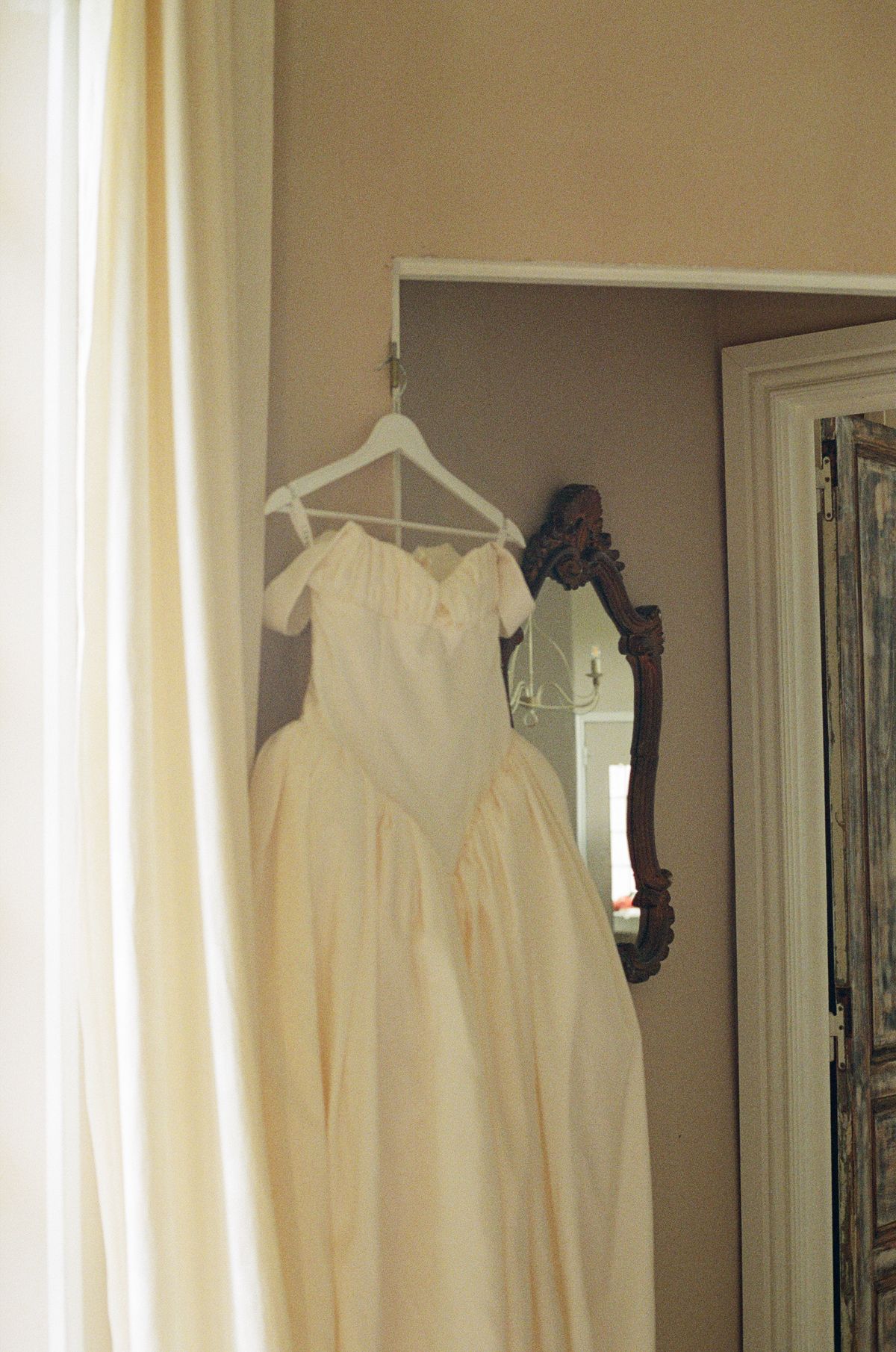 A white wedding dress with a full skirt hangs on a hanger beside a cream curtain in a softly lit room.