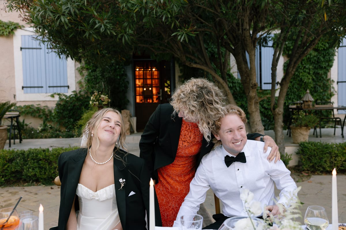 The bride and groom sit at an outdoor dinner table while a guest in an orange dress leans over and hugs them.