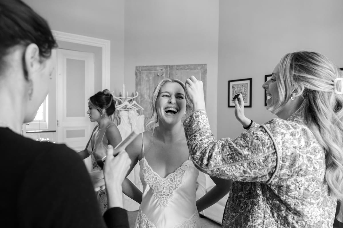 In black and white, a laughing bride in a slip dress has her hair adjusted by a woman in a patterned jacket.
