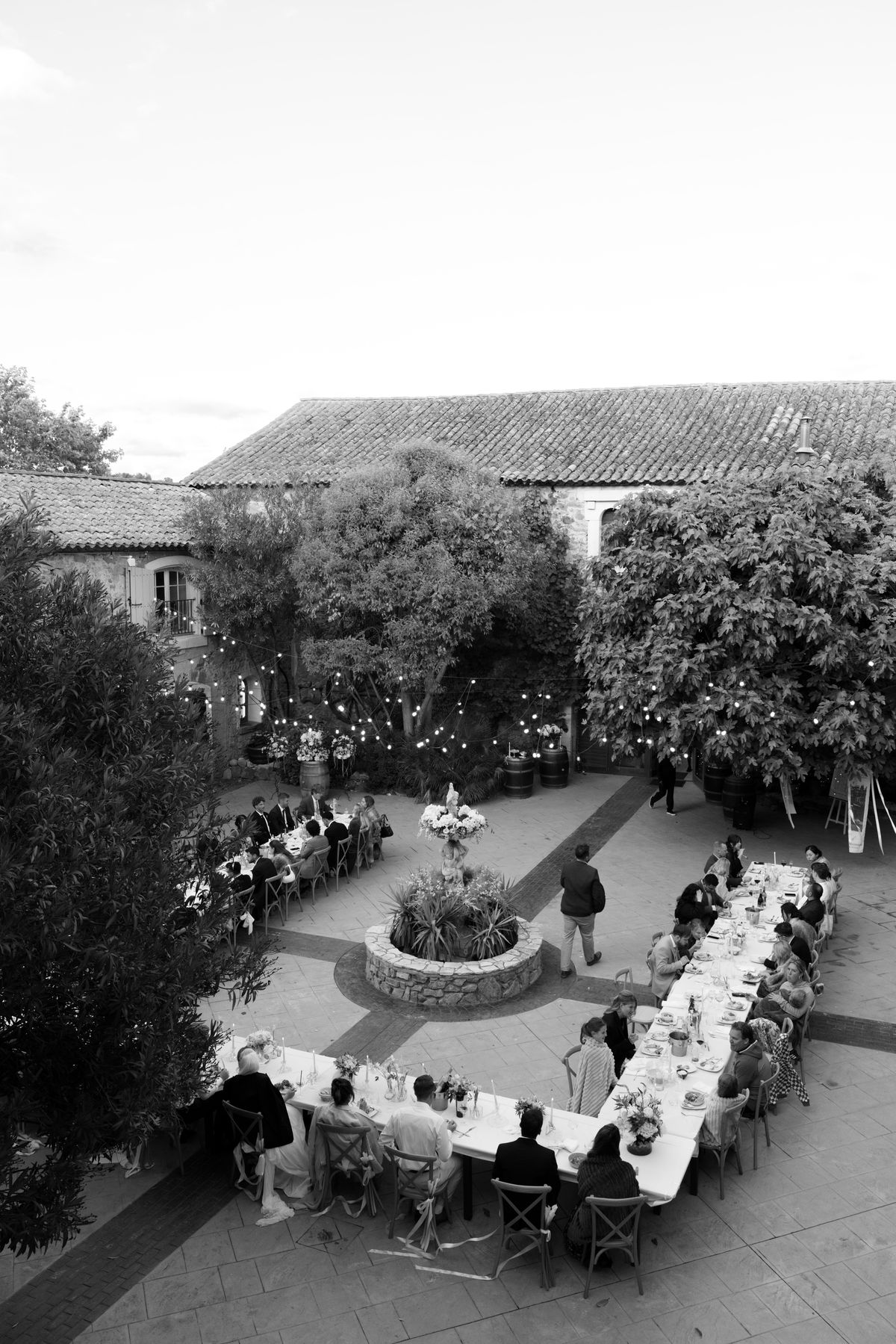 In black and white, guests sit at long tables arranged around a fountain in a courtyard, seen from above.
