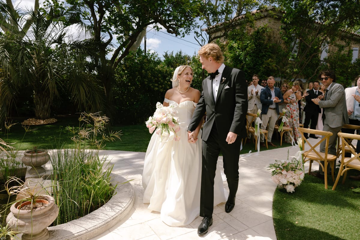 The bride holding a bouquet and the groom in a tuxedo walk hand in hand along a garden aisle, with guests standing behind them.