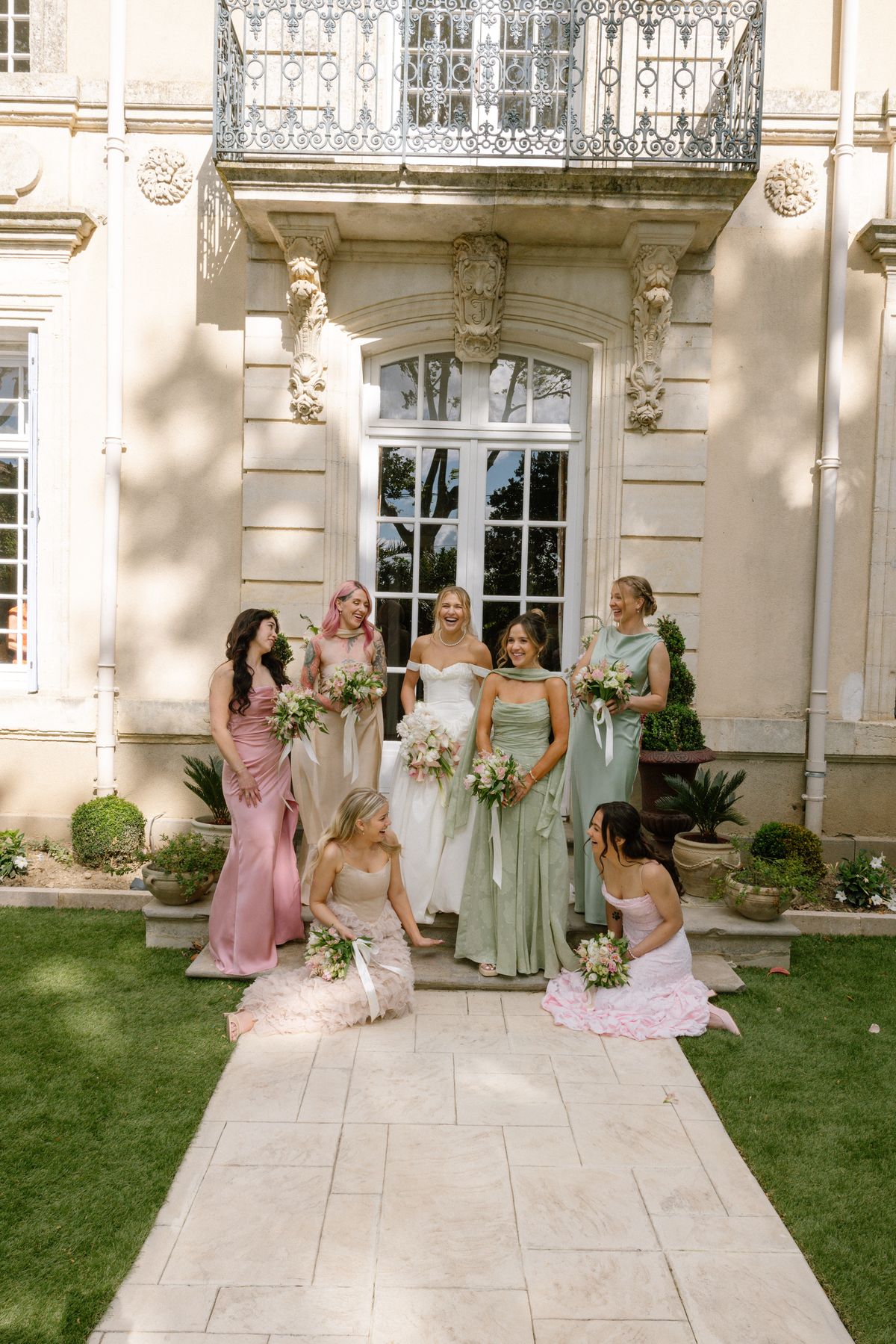 The bride and bridesmaids in pastel pink and green dresses pose with bouquets on the steps in front of a pale stone chateau.