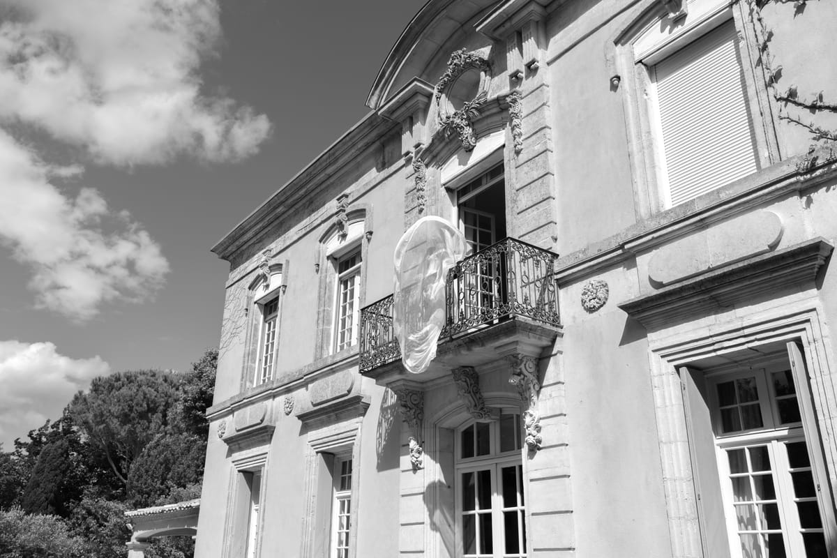 In black and white, the stone facade of a chateau with tall windows and a small balcony, under a sky with clouds.