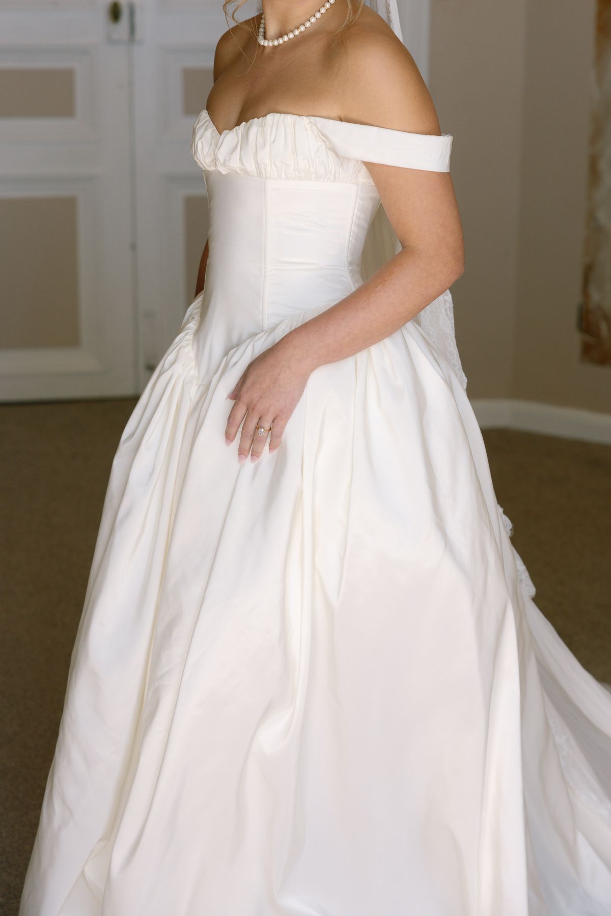 The bride stands in a pale room wearing an off-the-shoulder white gown with a full skirt and a pearl necklace, her face out of frame.