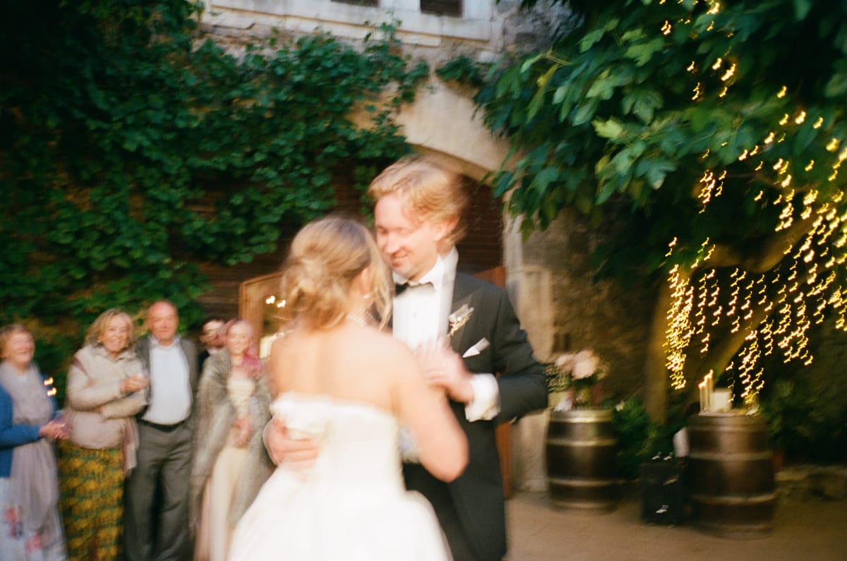 The couple dance together in a courtyard lit by string lights while guests look on in the background.