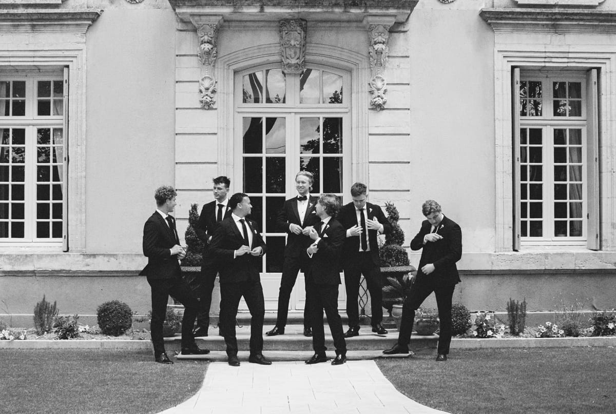 A group of men in dark suits stand and laugh on the steps in front of a grand stone building with tall windows.
