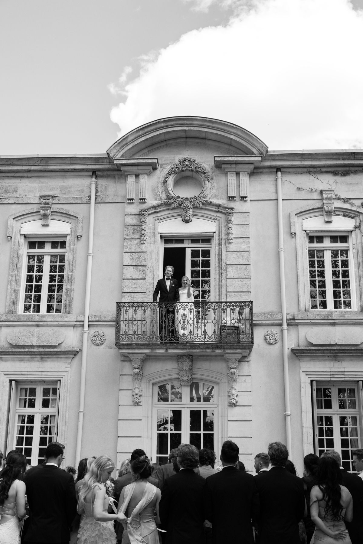 The couple stand on a wrought-iron balcony of a grand stone building while guests watch from below, seen from behind.