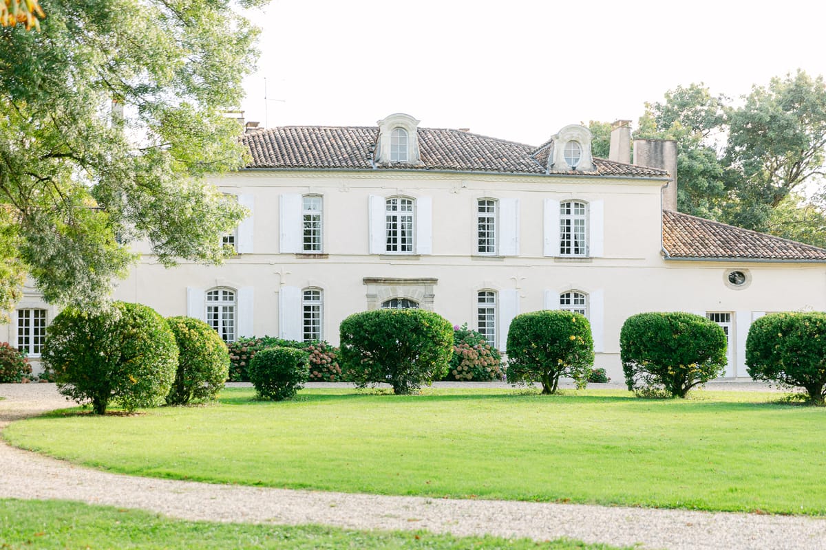 A pale stone chateau with shuttered windows stands behind a neat green lawn, rounded hedges and a gravel path, framed by large trees.