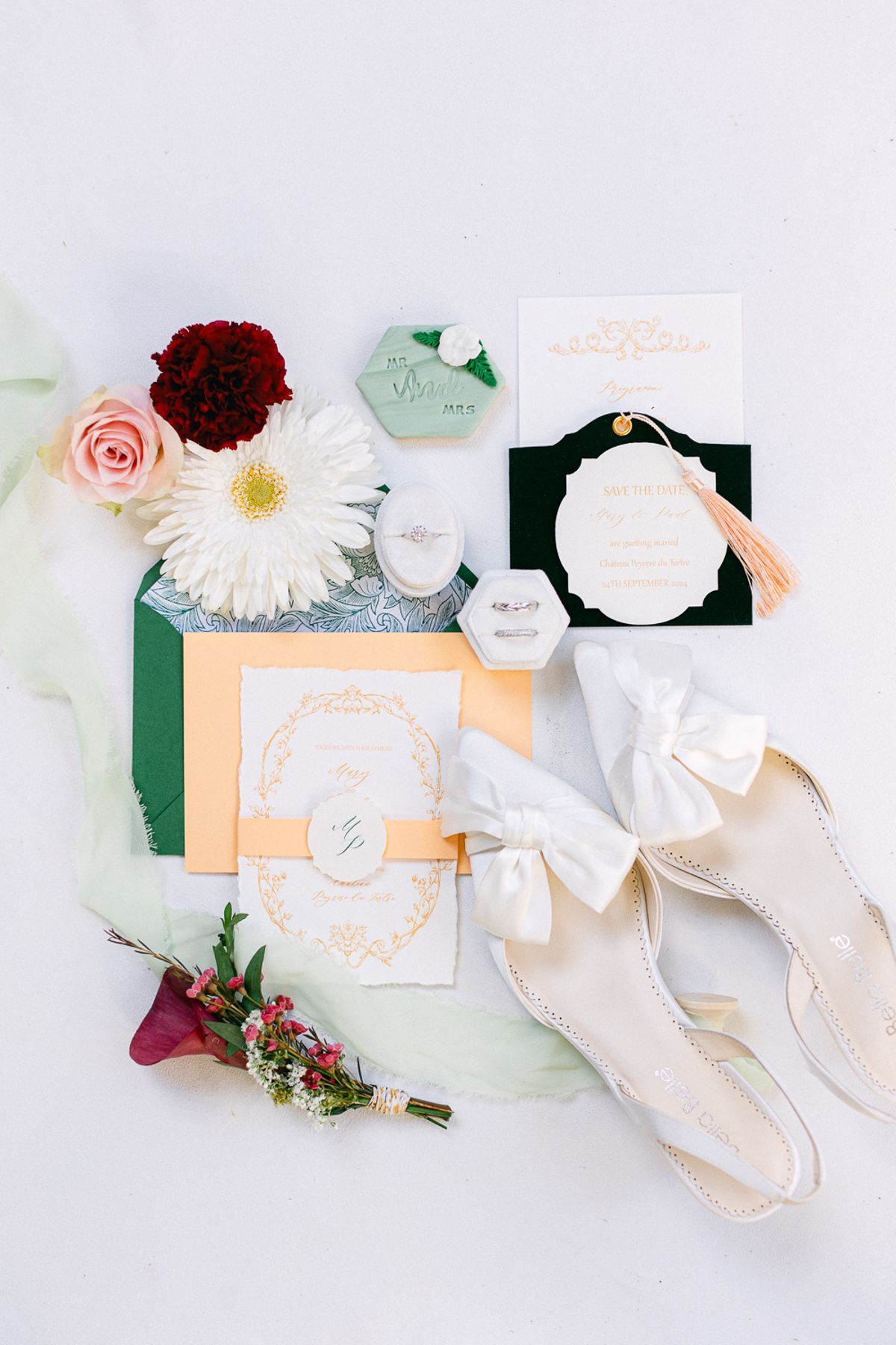 Flat lay of invitations, cream bow shoes, ribbons and red, pink and white flowers on a pale background.