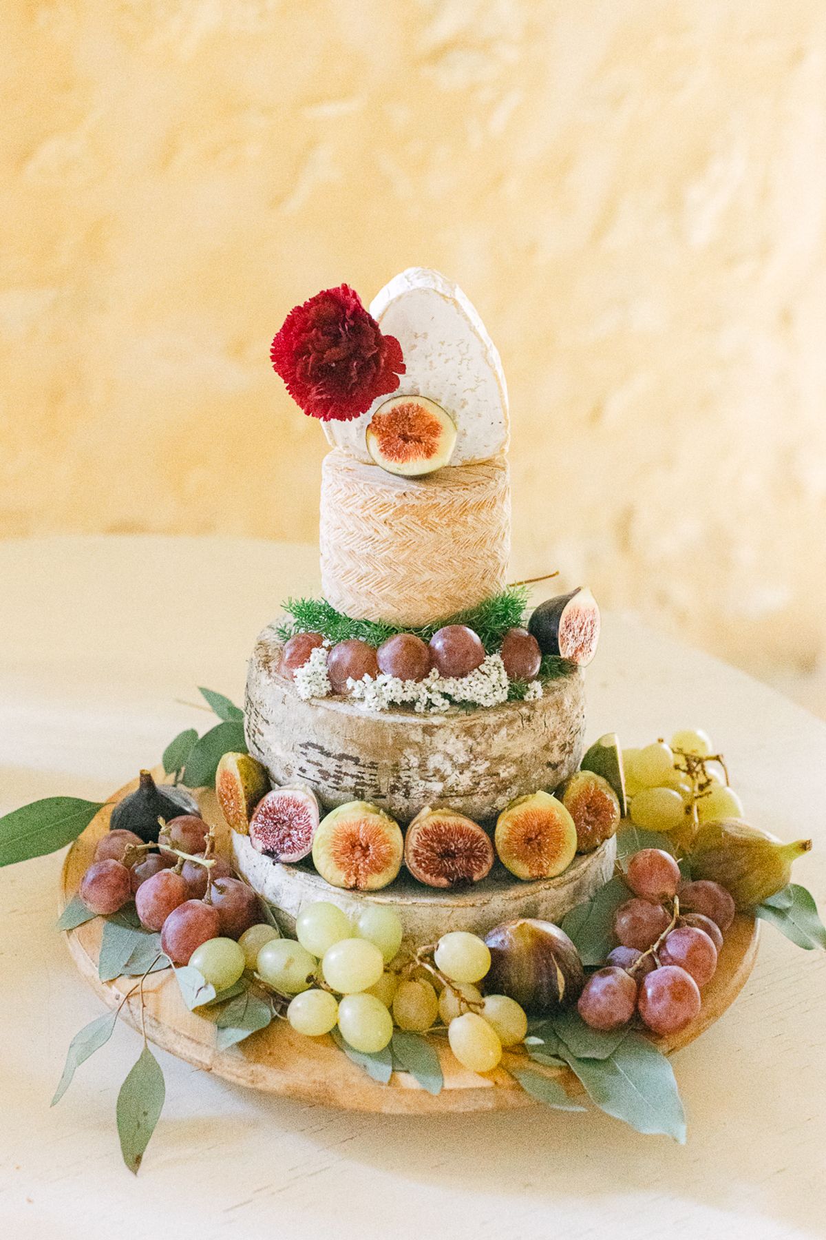 A tiered cheese cake topped with a red flower, decorated with figs, grapes and leaves on a wooden board.