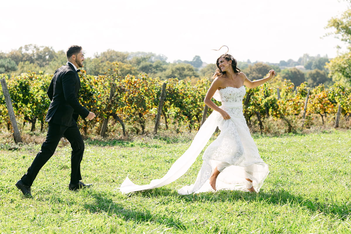 The groom in a dark suit strides across grass beside the bride in a white gown, with vineyards behind them.