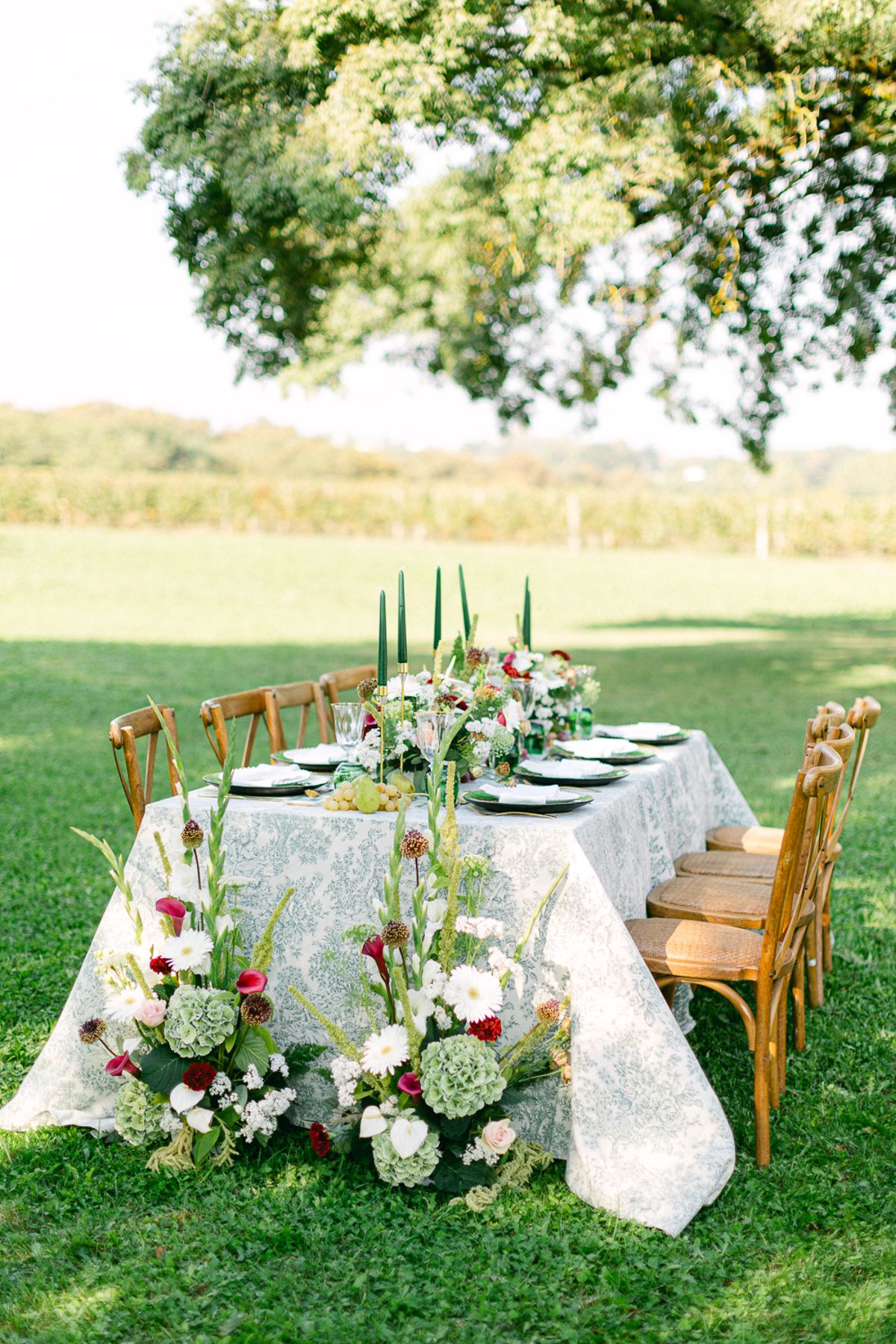 A long dining table with patterned linen, wooden chairs and tall floral arrangements set on a lawn beneath a large tree.
