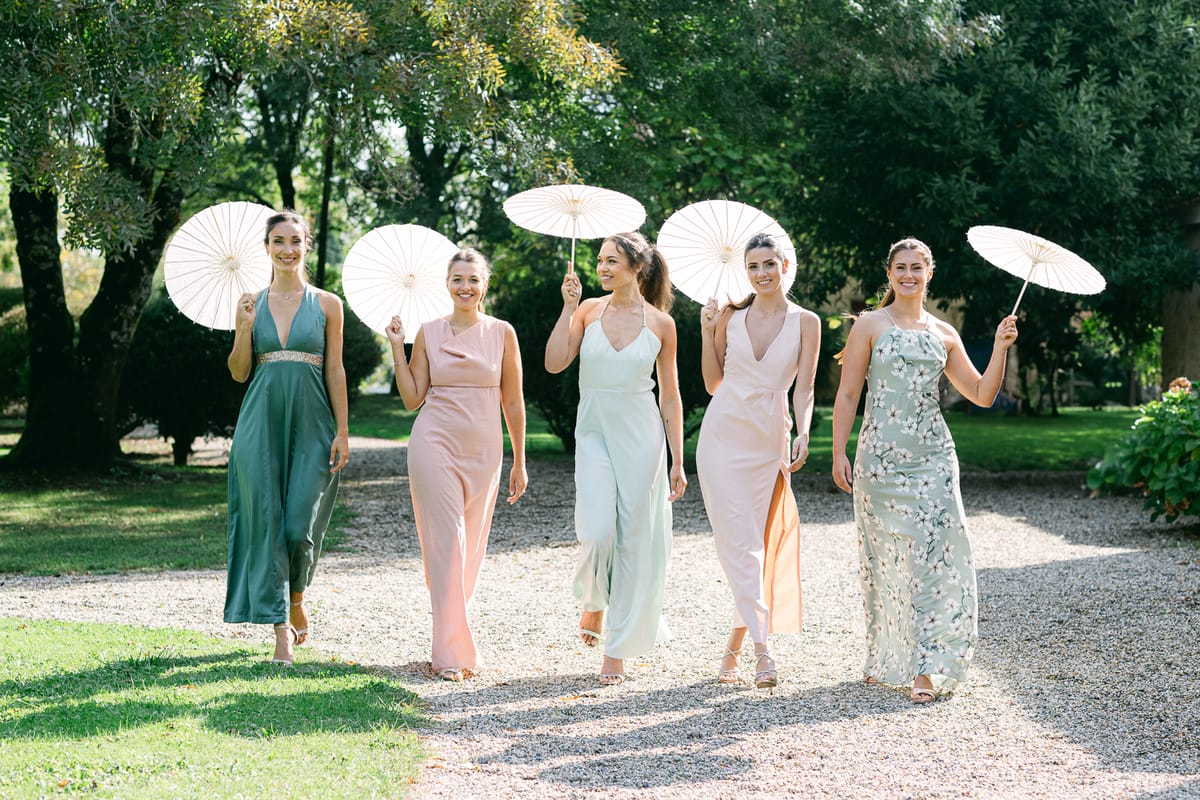 Five bridesmaids in pastel and green dresses walk along a gravel path under trees, each holding a white paper parasol.