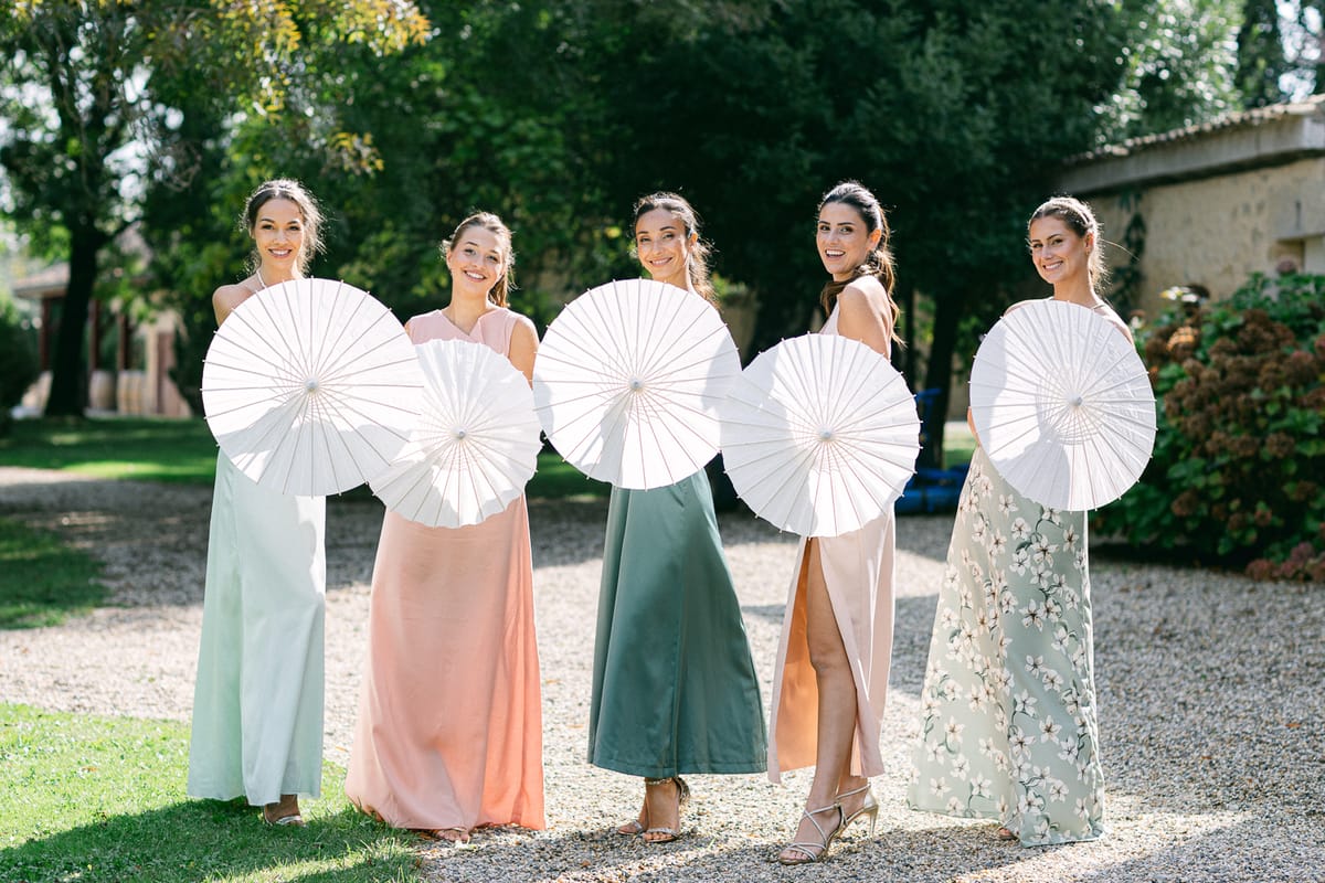 Five bridesmaids in pastel and green gowns stand in a row on gravel, holding white parasols in front of their bodies.