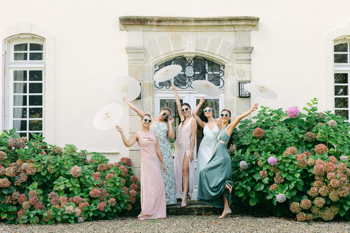 Bridesmaids in pastel dresses raise their arms and parasols in a chateau doorway, flanked by large pink hydrangea bushes.