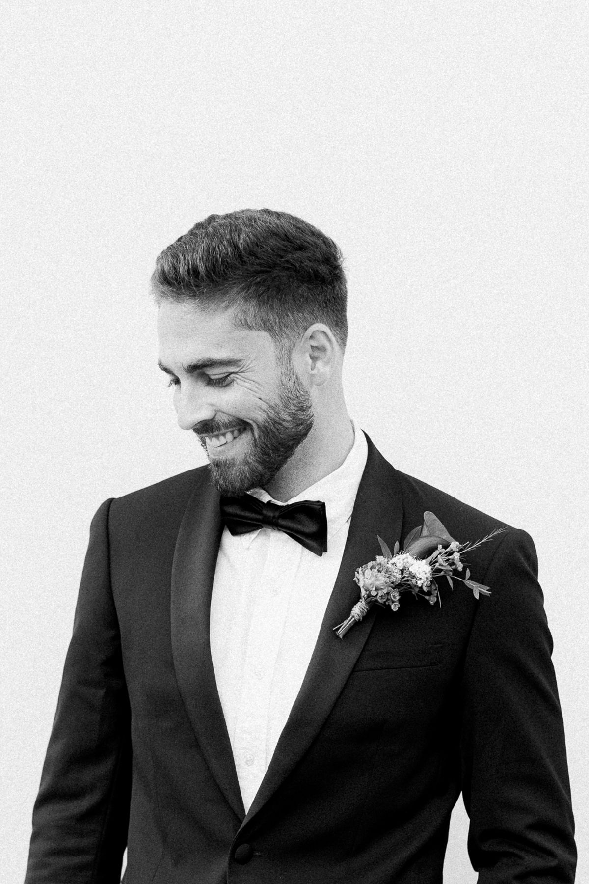 A black and white portrait of the groom in a tuxedo and bow tie, smiling and looking down, with a buttonhole flower.
