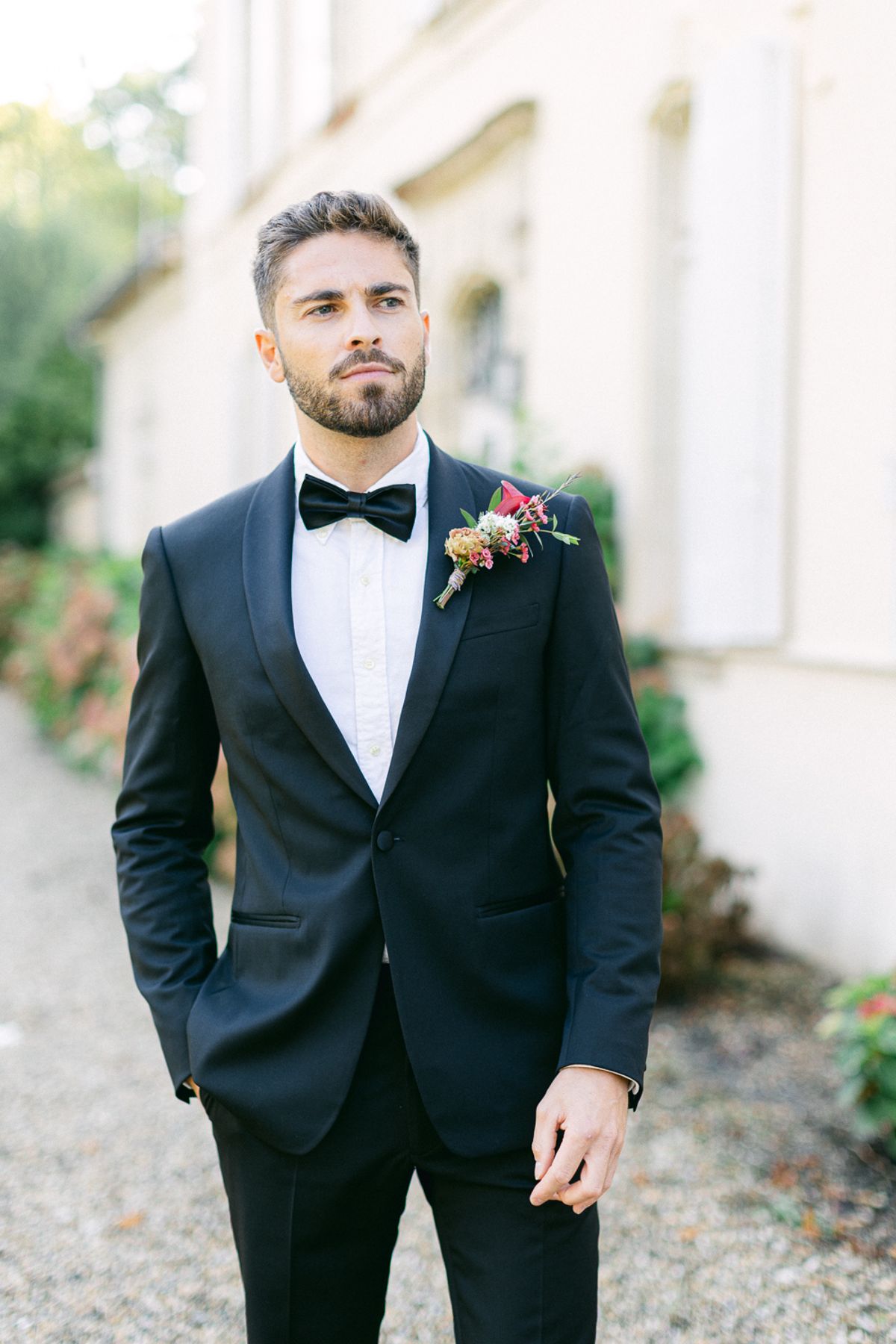The groom in a black tuxedo and bow tie stands with one hand in his pocket on a gravel path beside a pale building.