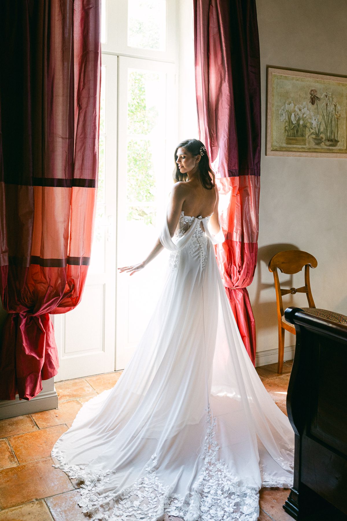 The bride, seen from behind in a lace gown with a long train, stands at a tall window framed by red curtains.