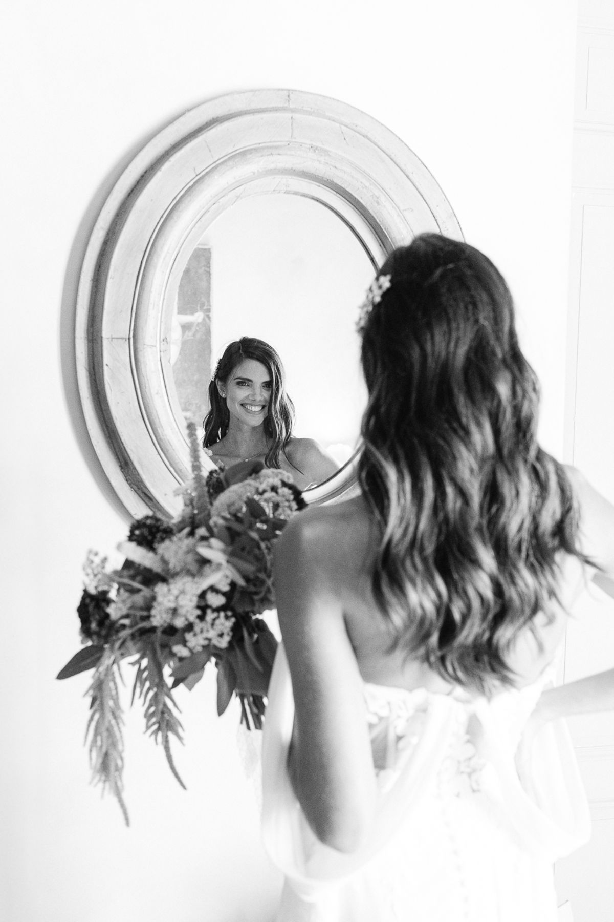 A black and white image of the bride from behind, holding a bouquet and looking at her reflection in a round mirror.