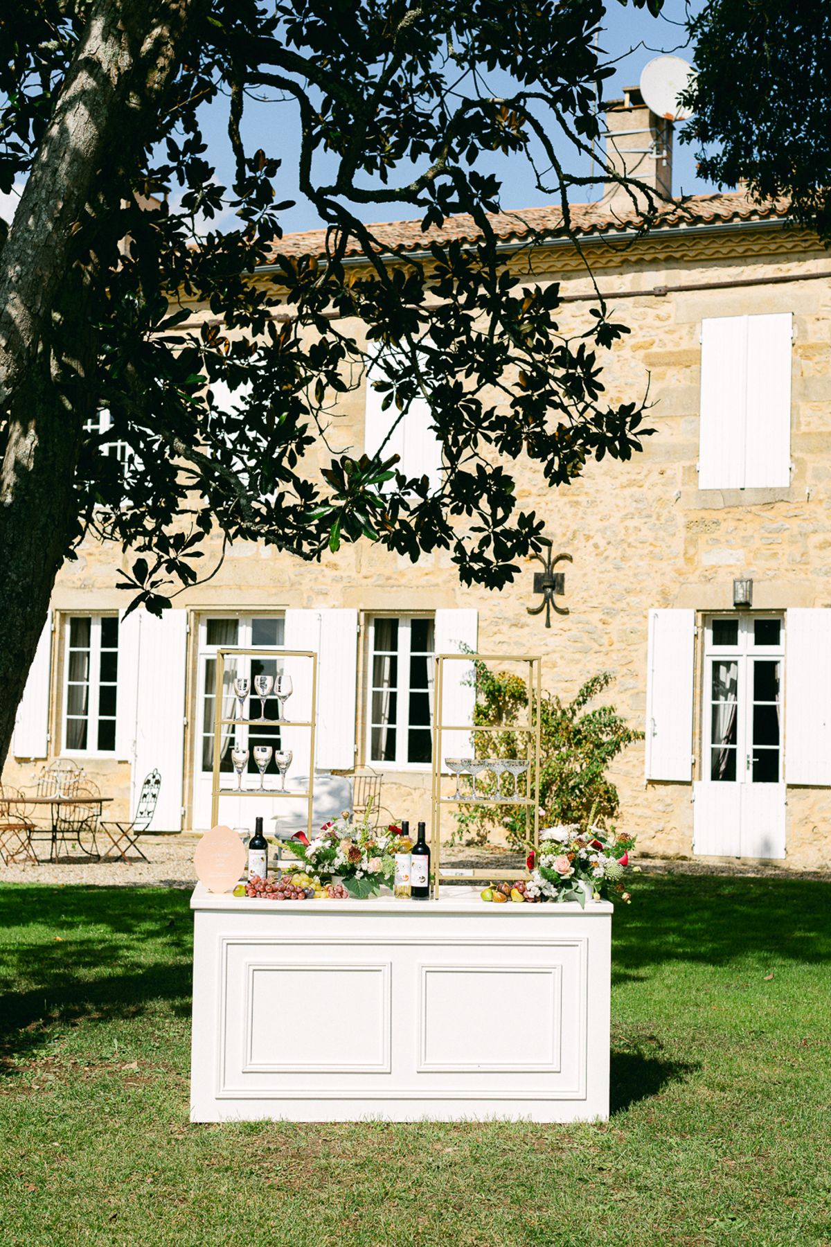 A white cabinet holding bottles and flowers stands on a lawn in front of a stone house with white shutters.