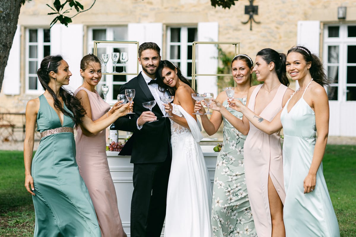 The couple and five bridesmaids in green and pink dresses raise glasses together on a lawn in front of a stone house.