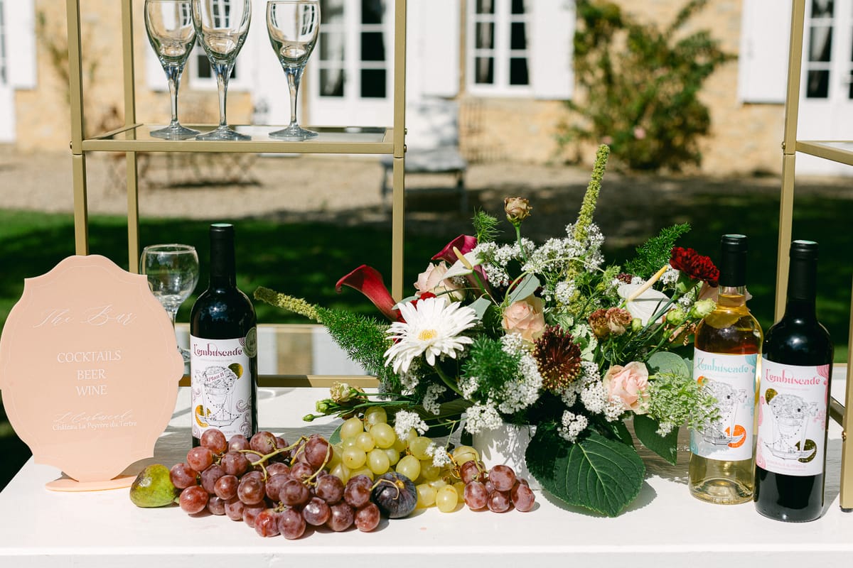 A drinks table with wine bottles, bunches of grapes, a floral arrangement, a peach sign and champagne glasses on a gold stand.