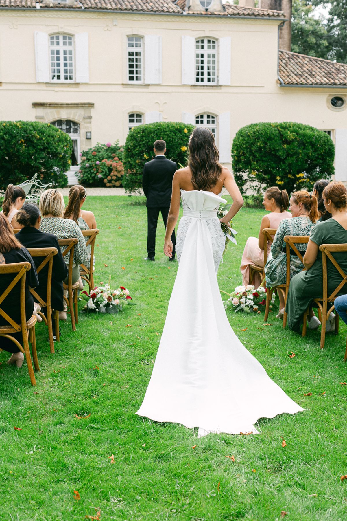 The bride in a long white gown walks down a grass aisle between seated guests towards a figure waiting before a stone house.