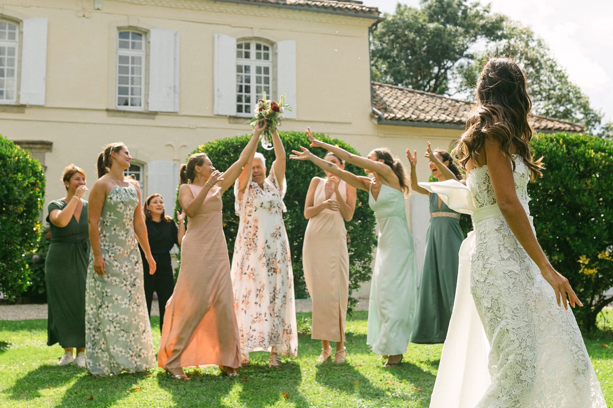 The bride in a white gown tosses her bouquet towards bridesmaids with raised arms on a lawn in front of a chateau.
